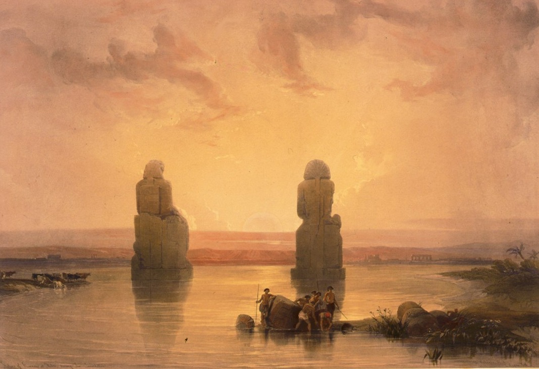 Two colossal seated statues standing in floodwater at sunset, with cattle wading in the shallows and men at work beside a fallen block