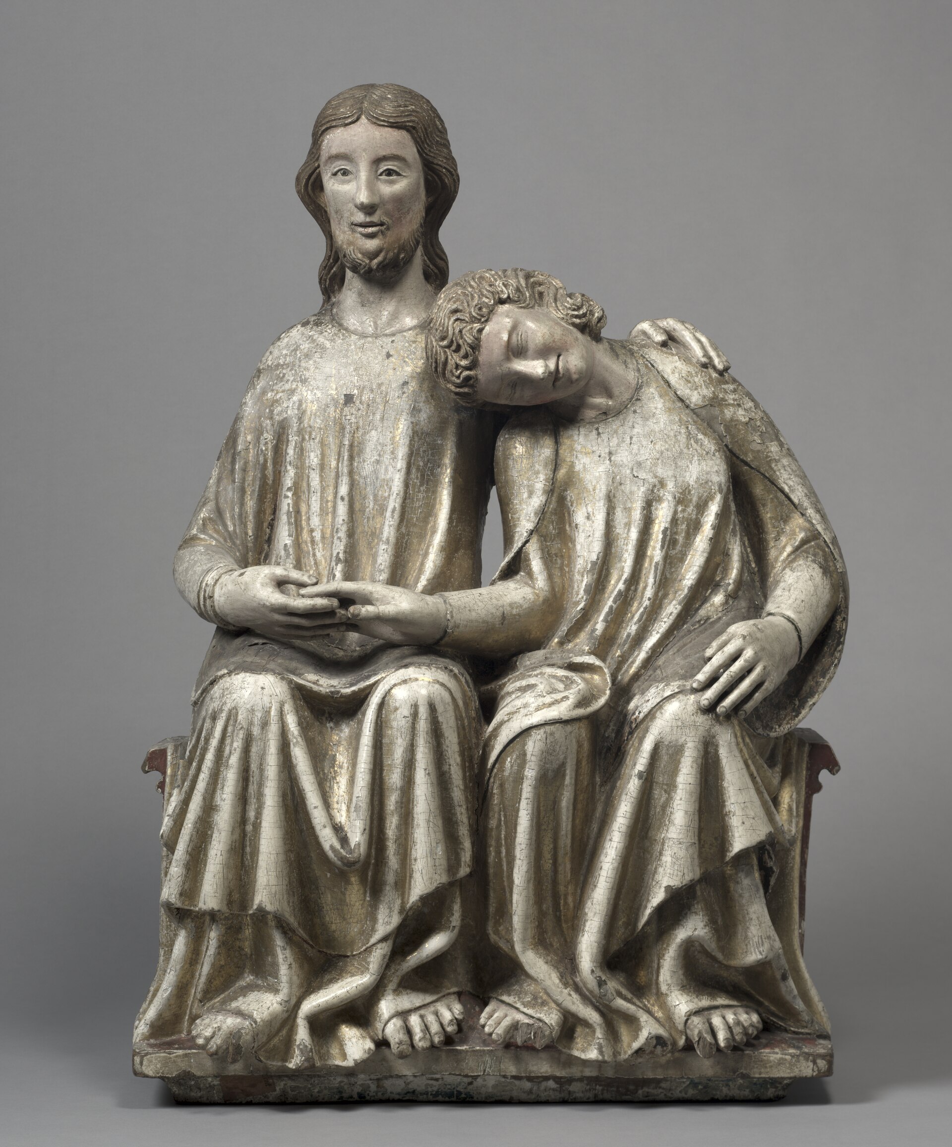 A painted and gilded wooden sculpture of Christ seated beside the young apostle John, who rests his head on Christ's shoulder with eyes closed while Christ holds his hand and wraps an arm around him