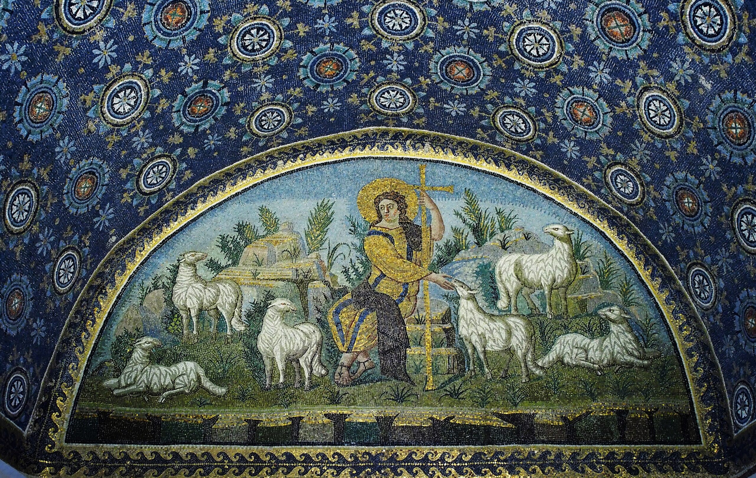 A golden lunette mosaic of Christ as the Good Shepherd, seated among six white sheep in a green pasture, holding a tall gold cross and reaching out to feed one of the flock, beneath a deep blue vault of starry rosettes