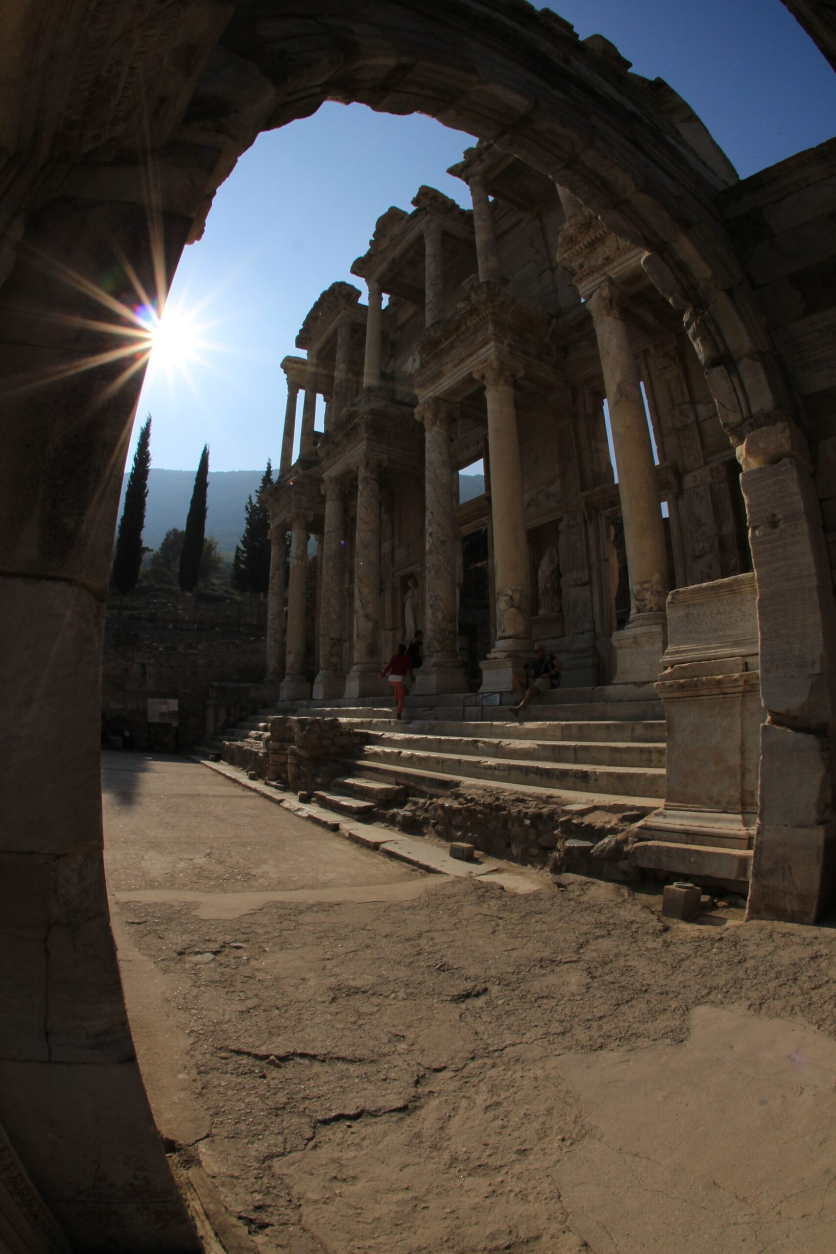 The columned marble facade of the Library of Celsus in Ephesus, seen through a ruined stone archway against a bright sun