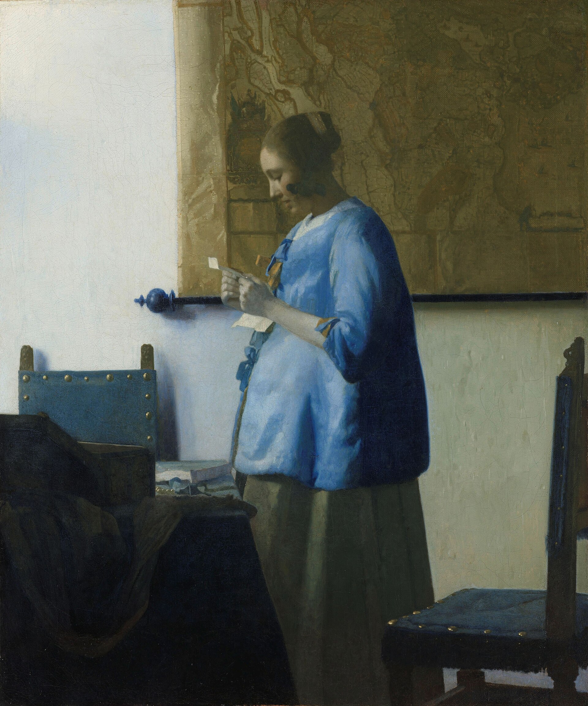 A young woman in a blue jacket stands by a window in a quiet room, absorbed in reading a letter held close in both hands, a large map hanging on the wall behind her