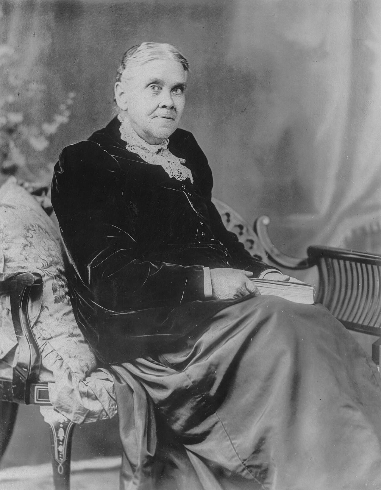 An elderly Ellen G. White with her hair parted in the middle, wearing a dark velvet jacket and lace collar, seated in an armchair with a book resting on her lap