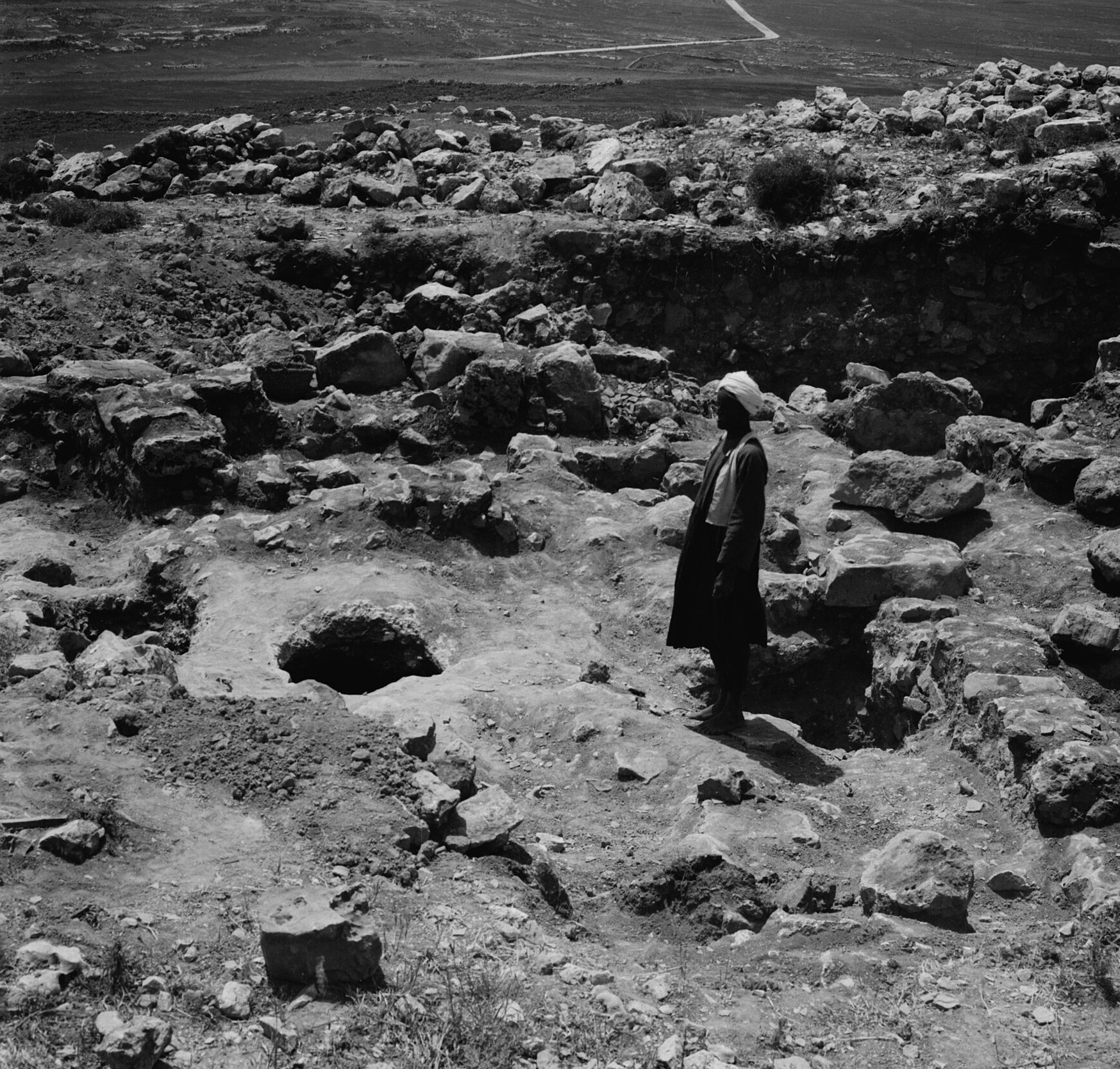 The Dungeon And The Ethiopian 1 Old black-and-white photograph of an excavated hilltop of tumbled stones, with the round dark mouth of an ancient cistern cut into the rock and a man standing beside it