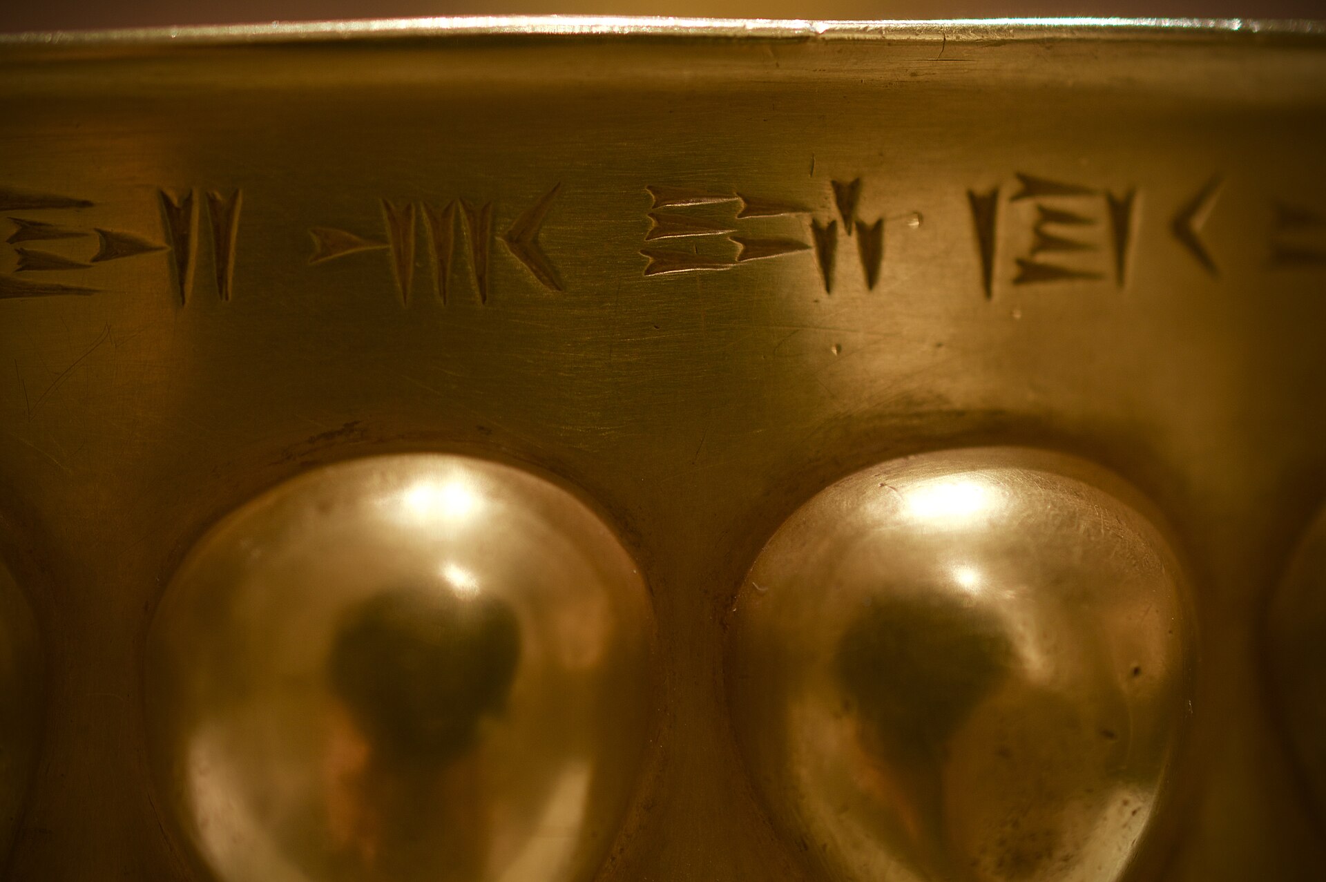 How Is The Gold Become Dim 1 Close-up of the rim of an ancient gold bowl, with a line of wedge-shaped cuneiform writing above a row of polished, rounded flutes that reflect the light